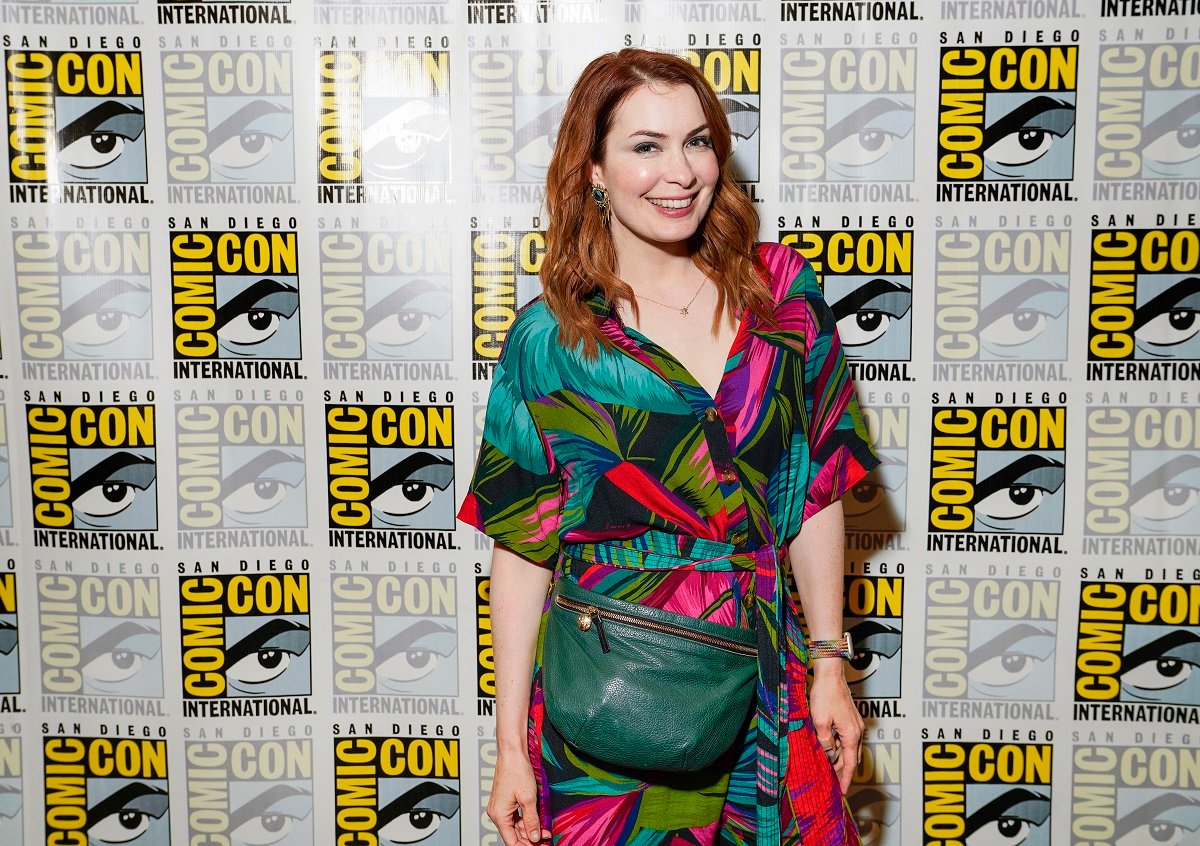 Felicia Day (white woman with long red hair wearing a plant print dress and a green purse) standing in front of a San Diego Comic-Con step and repeat, smiling. 