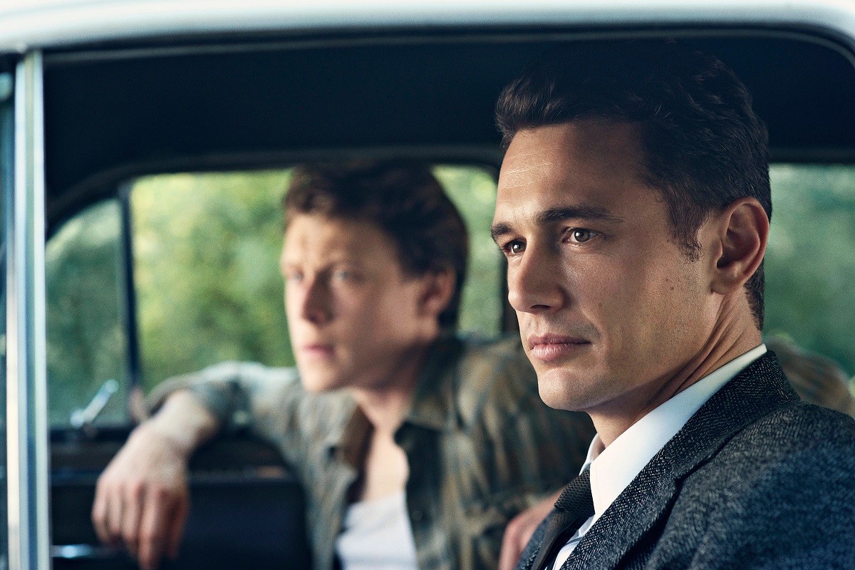 James Franco as Jake Epping in a scene from Hulu's '11.22.63.' He is a white man with short, dark hair wearing a suit sitting in the driver's seat of a 1960s car looking out his window. There is a white man in the passenger's seat out of focus. 