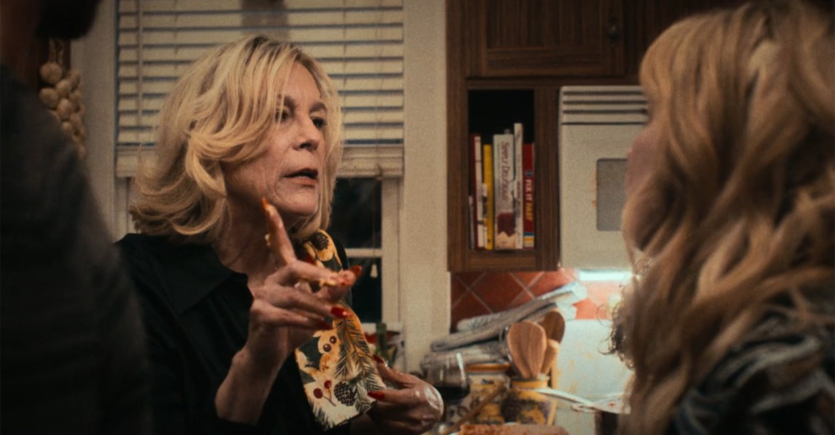 A blonde woman in a hectic home kitchen giving directions to those who walk in.