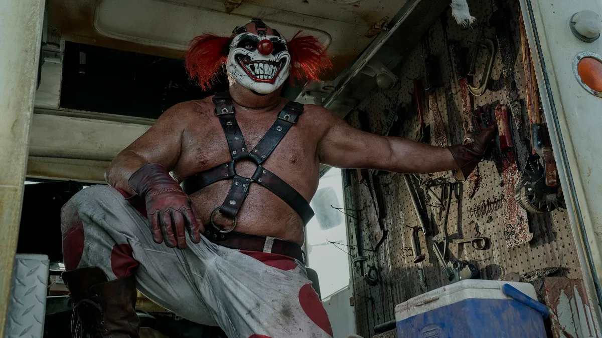 Joe Seanoa (large, muscular white wrestler-looking man) as Sweet Tooth in Peacock's 'Twisted Metal.' He is wearing a clown mask, a leather harness over a bare chest, red gloves, and white pants with large red polka dots on them He's standing in the back of a truck with one foot propped up on something and one hand propping him up against a wall with a bunch of sharp tools hanging on it. He's looking out of the open back of the truck.