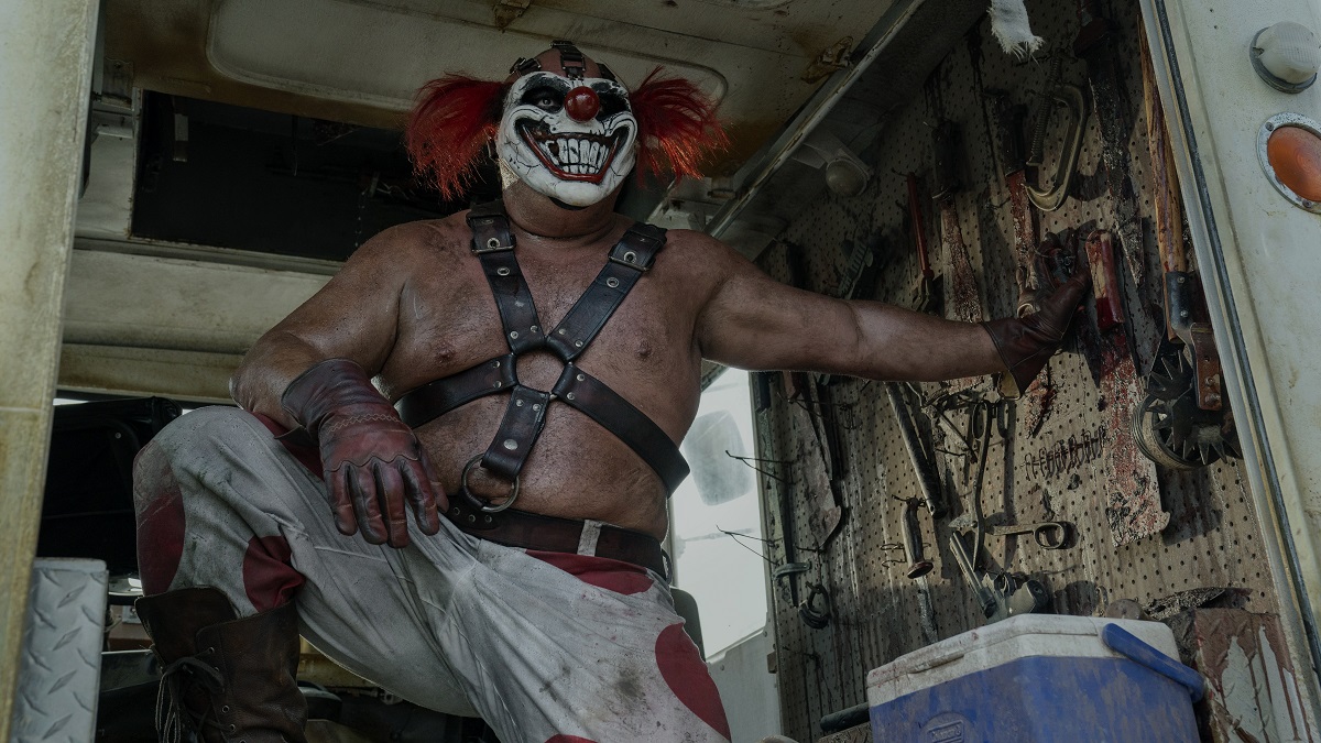 Joe Seanoa (large, muscular white wrestler-looking man) as Sweet Tooth in Peacock's 'Twisted Metal.' He is wearing a clown mask, a leather harness over a bare chest, red gloves, and white pants with large red polka dots on them He's standing in the back of a truck with one foot propped up on something and one hand propping him up against a wall with a bunch of sharp tools hanging on it. He's looking out of the open back of the truck.