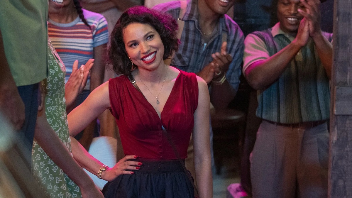 Jurnee Smollett as Leti in a scene from HBO's 'Lovecraft Country.' She is a light-skinned Black woman with wavy dark chin-length hair wearing hoop earrings, a red sleeveless blouse and black pants as she stands smiling with her hand on her hip in the middle of a party. 