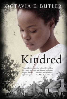 Book cover of Octavia E. Butler's novel, 'Kindred.' Butler's name is at the top in white caps. A Black woman with cropped, natural hair with a downward gaze in a white blouse takes up most of the cover. At the bottom, we see a row of houses in black-and-white. The title is in the middle of the book in black letters, and beneath it is a quote by Harlan Ellison that reads "Octavia Butler is a writer who will be with us for a long, long time, and Kindred is that rare magical artifact...the novel one returns to again and again."