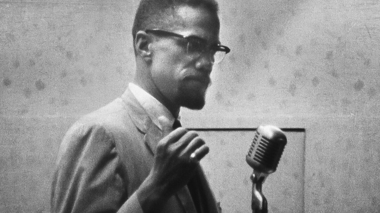 Archival footage of Malcolm X speaking into a microphone in 'Who Killed Malcolm X?'
