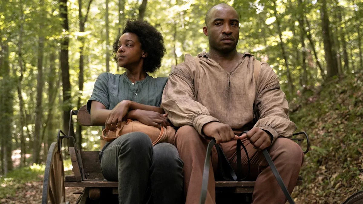 Mallori Johnson as Dana and Austin Smith as Luke in a scene from Hulu's 'Kindred.' They are both Black and seated in a wagon with Luke holding the reins. Dana has her natural hair in an afro and is wearing a blue-grey shirt with buttons at the collar and dark pants. Luke is bald and wearing a pinkish-grey long-sleeved shirt and orange-ish pants. They are riding through the woods. 