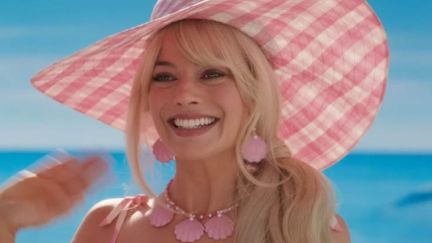 Image of Margot Robbie as Barbie in the Warner Bros. film, 'Barbie.' It's a close-up of Barbie, a white woman with long, blonde hair wearing a pink and white checkered wide-brimmed hat, large pink seashell earrings, and a matching pink seashell necklace. Her hair is in a ponytail pulled over her shoulder with long strands of hair flying free. She's smiling and waving.