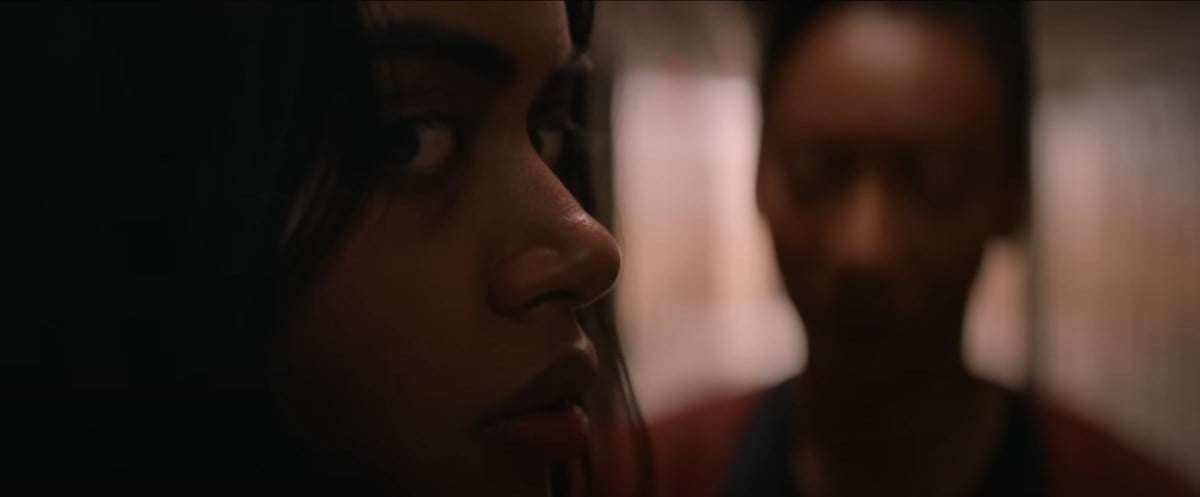 Megan Suri as Samidha (aka "Sam") in a scene from the NEON horror feature, 'It Lives Inside.' Samidha is an Indian-American teenage girl with long dark hair. We see her close-up and in profile as she looks over her shoulder warily at something approaching. Her teacher, played by Betty Gabriel (a Black woman with her hair up in a bun) stands with her in a school hallway out of focus.