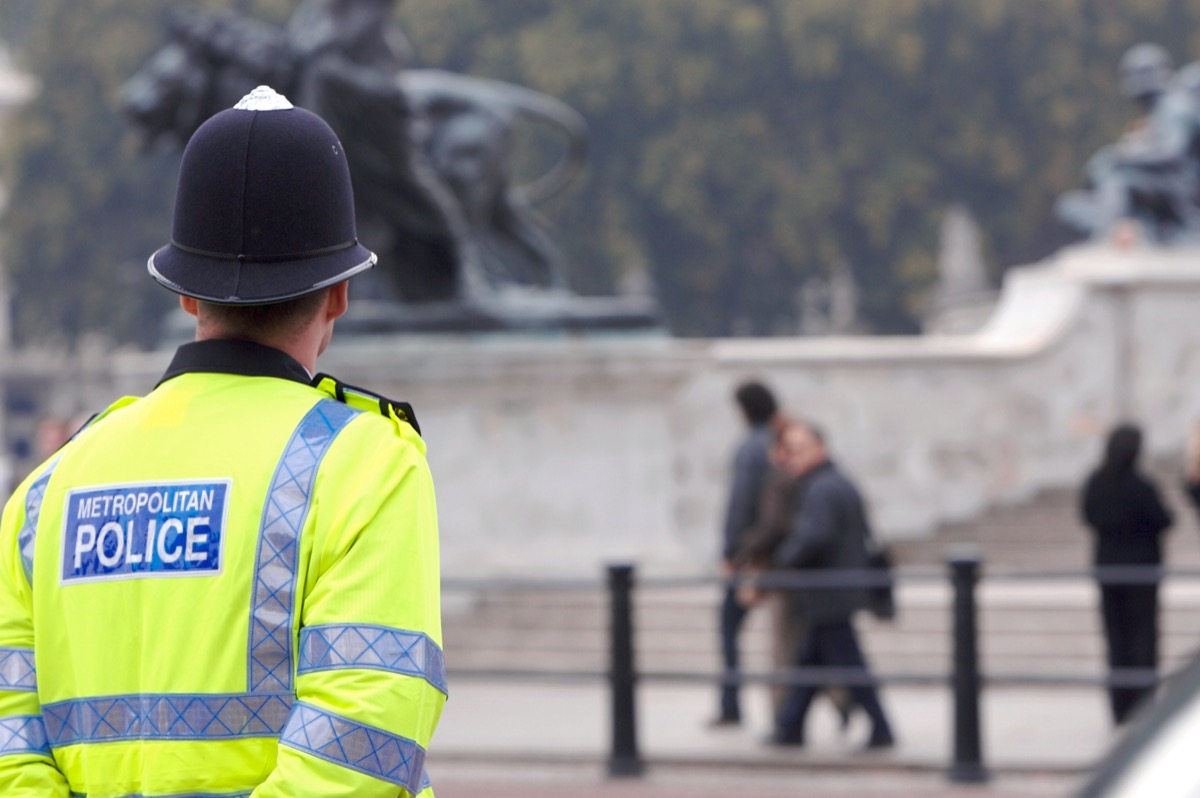 A British police man stands on duty across the road from Buckingham Palace, as tourists pass by in the background on their way to see the Changing of the Guard.
