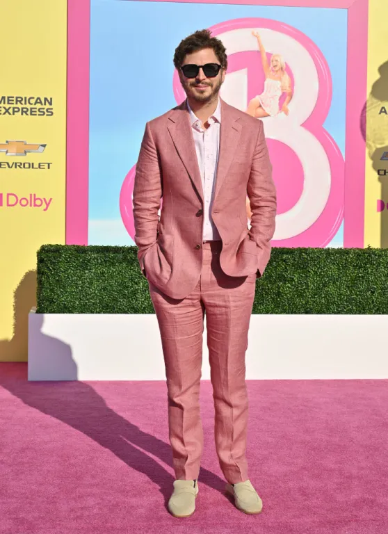 Michael Cera wears a light pink suit, white shirt, and white shoes. He stands on a pink carpet with the Barbie logo behind him.