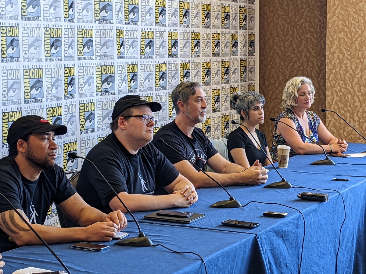 Zeke Alton (Black man with a beard wearing a black baseball cap and black t-shirt), Duncan Crabtree-Ireland (white man with glasses wearing a black baseball cap and black t-shirt), Tim Friedlander (white man with salt-and-pepper beard and hair wearing a black t-shirt), Ashly Burch (white woman with salt-and-pepper hair pulled into a bun wearing a black scoop necked shirt), and Cissy Jones (white woman with chin-length wavy blonde hair wearing a short-sleeved floral print dress) sit at a dais in front of microphones at SDCC.