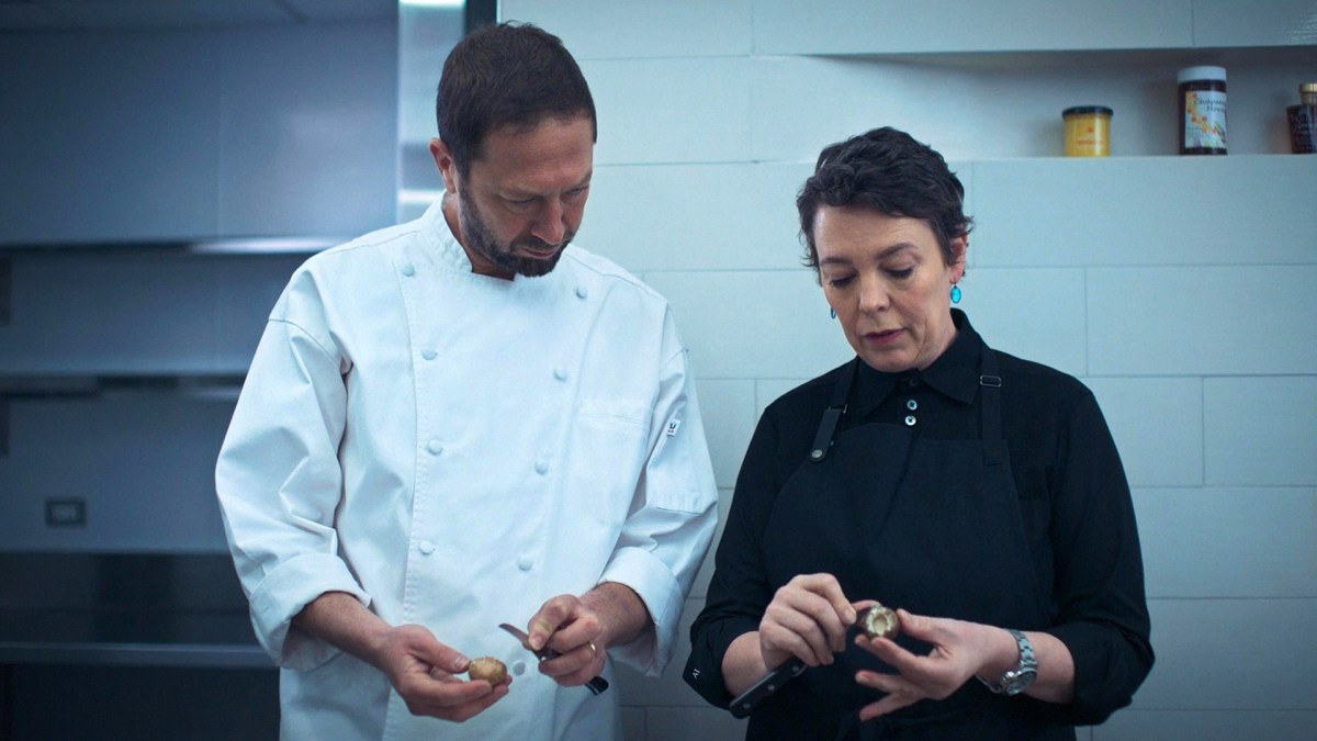 Two people stand in a stark white kitchen peeling mushrooms while in conversation.