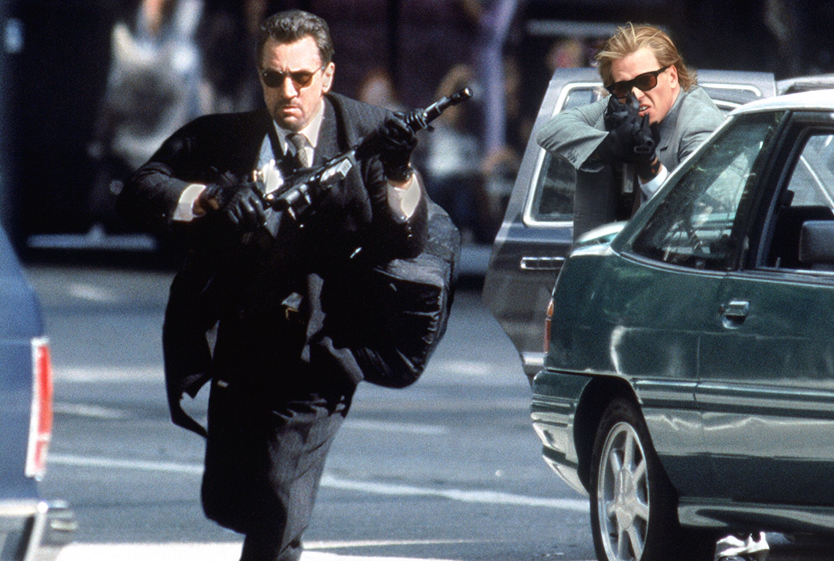 A man in a suit and sunglasses runs through the street with a gun while another man in a car points a gun in the same direction in "Heat" 