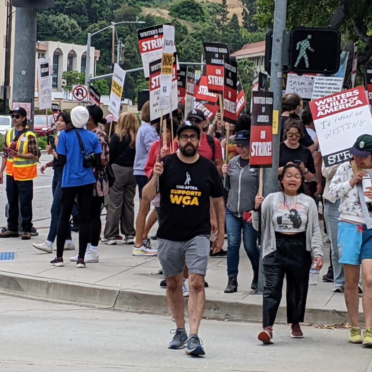 Image of a crowd of people crossing the street holding Writers Guild of America picket signs with one white man with a dark beard, glasses, wearing shorts, a black t-shirt, and a baseball cap is in the foreground. His shirt reads "SAG-AFTRA Supports WGA" and he's holding a SAG-AFTRA strike sign.