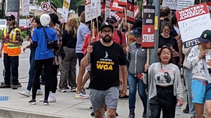 Image of a crowd of people crossing the street holding Writers Guild of America picket signs with one white man with a dark beard, glasses, wearing shorts, a black t-shirt, and a baseball cap is in the foreground. His shirt reads 