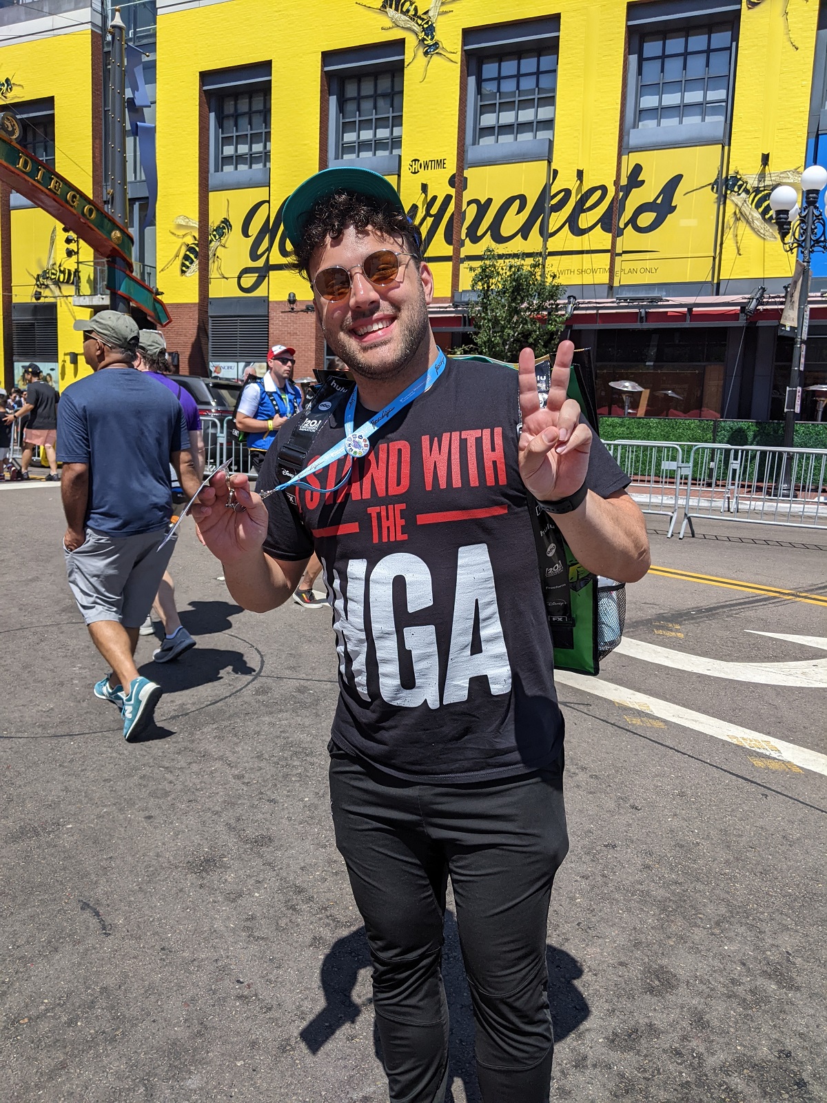 Image of a young, white man with dark curly hair wearing a baseball cap, sunglasses, a black t-shirt that reads "I STAND WITH THE WGA" and black pants. He's flashing a peace sign and smiling. 