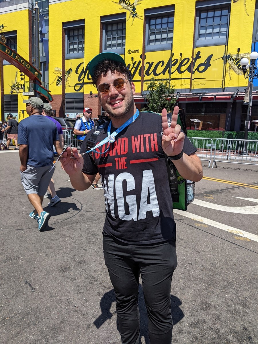 Image of a young, white man with dark curly hair wearing a baseball cap, sunglasses, a black t-shirt that reads "I STAND WITH THE WGA" and black pants. He's flashing a peace sign and smiling.
