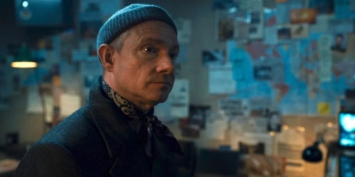 Agent Ross, wearing a black beanie, looks at someone with dozens of papers pinned to the wall behind him.