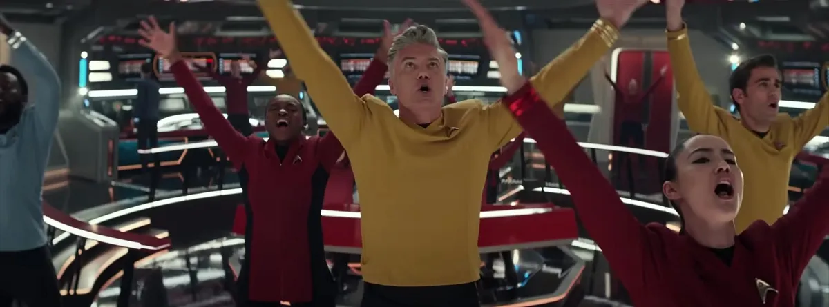 Screengrab from the 'Strange New Worlds' musical episode, "Subspace Rhapsody." Captain Pike is in the center of several crew members on the bridge, all of whom have their arms in the air, mid-song.