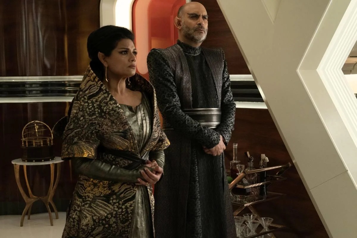 TPril (Ellora Patniak) and Sevet (Michael Benyaer) in a scene from 'Strange New Worlds.' They are both Vulcans with pointed ears. Patniak is a brown-skinned woman of Indian descent. T'Pril has her long, dark hair pulled up around her head, wears thin, dangling earrings, and is wearing a shiny, dark grey dress with a dark grey and gold high-necked cloak over it. Her hands are clasped. Benyaer is a brown-skinned man of Indian descent. Sevet is bald with a salt-and-pepper beard. He wears a long, long-sleeved, black and grey robe with a black and silver band around the middle. He stands with his hands folded in front of him.