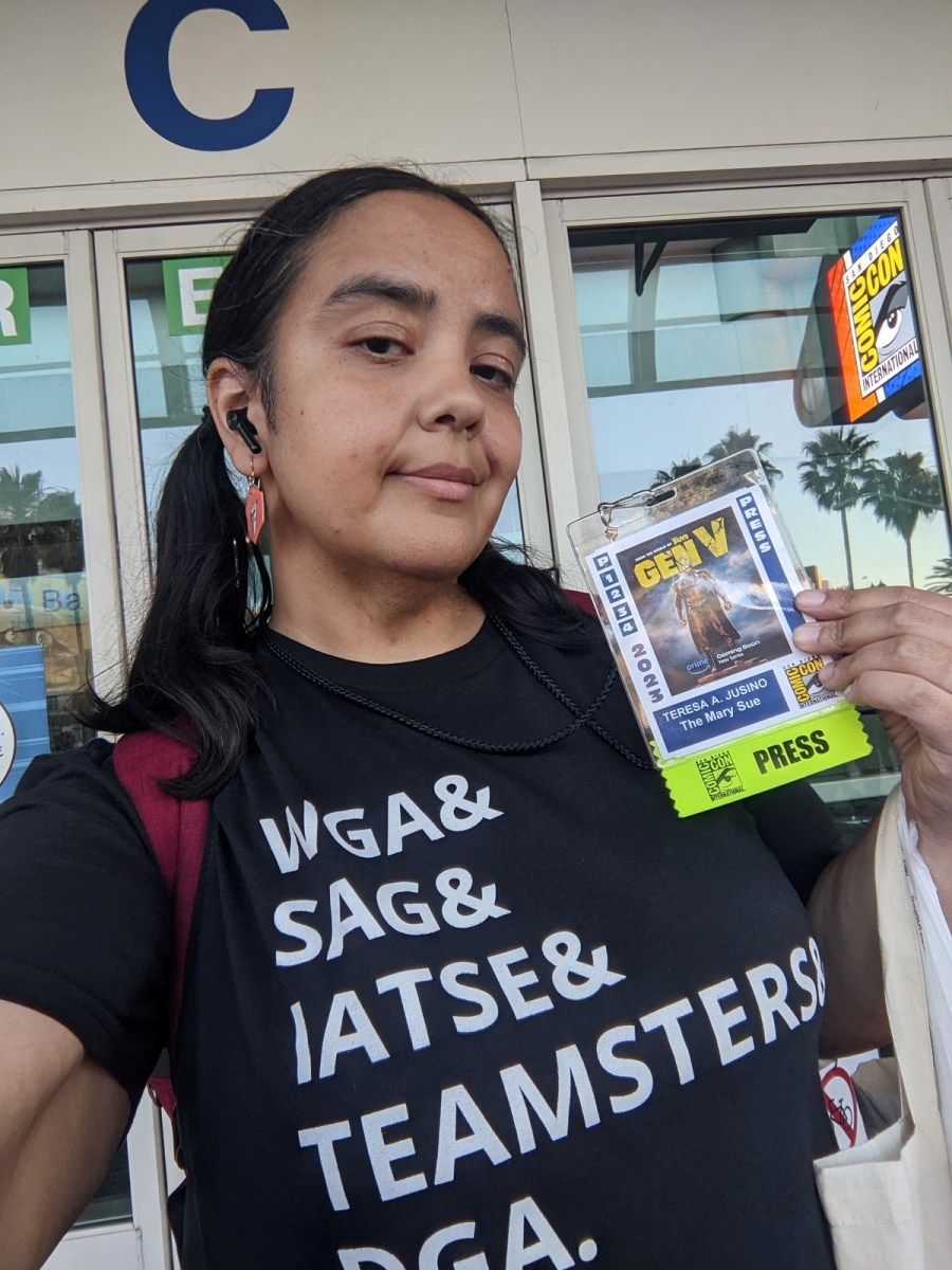 Image of Teresa Jusino (brown Latina with long, dark hair in pigtails) wearing red dangly earrings and a t-shirt that reads "WGA & SAG & IATSE & TEAMSTERS & DGA." She is holding up her Comic-Con badge which has her name and outlet on it, and a "Press" sticker attached to the bottom.