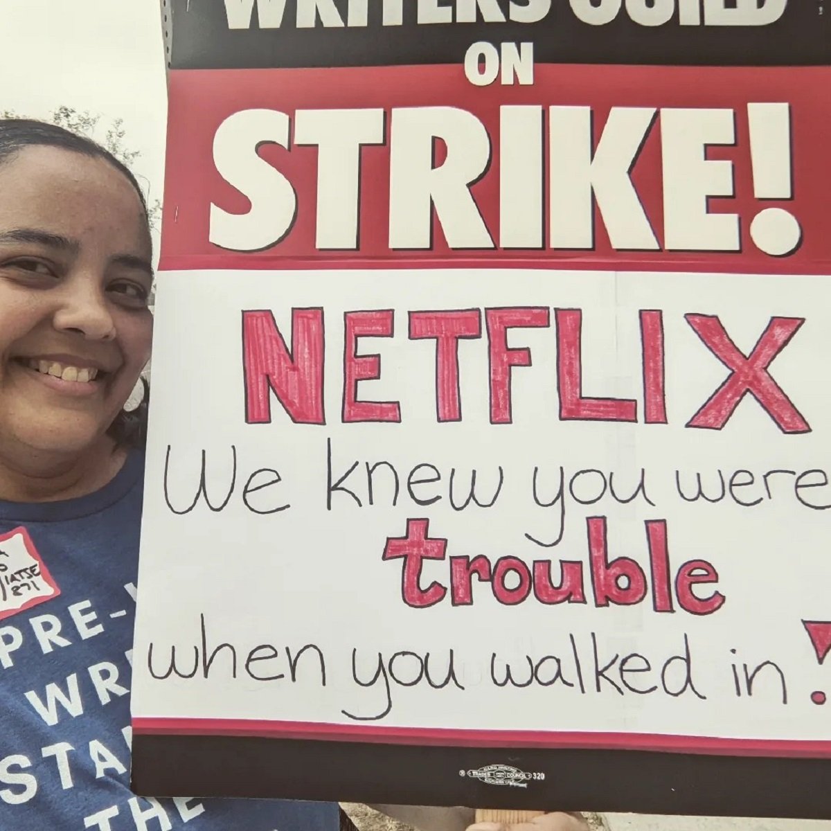 Square image of Teresa Jusino (a brown Latina with long, black hair in pigtails) holding a picket sign that reads "Writers Guild on Strike!" on top, and then "Netflix - we knew you were trouble when you walked in!" handwritten on the bottom.