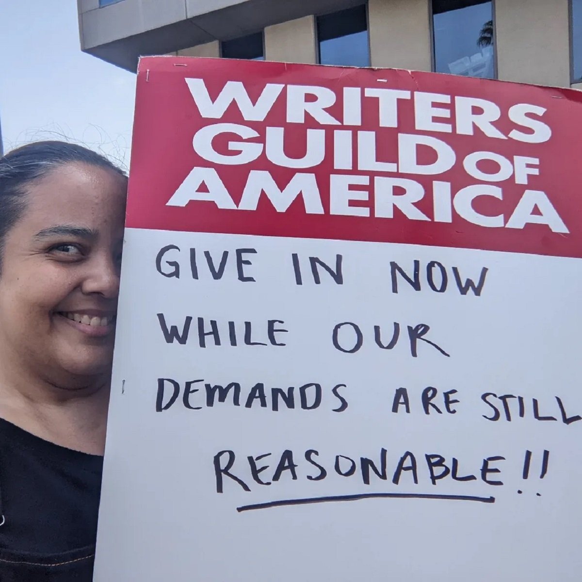 Square image of Teresa Jusino (a brown Latina with long, black hair in a ponytail) holding a picket sign that reads "Writers Guild of America" on top, and then "Give in now while our demands are still REASONABLE!" handwritten on the bottom.