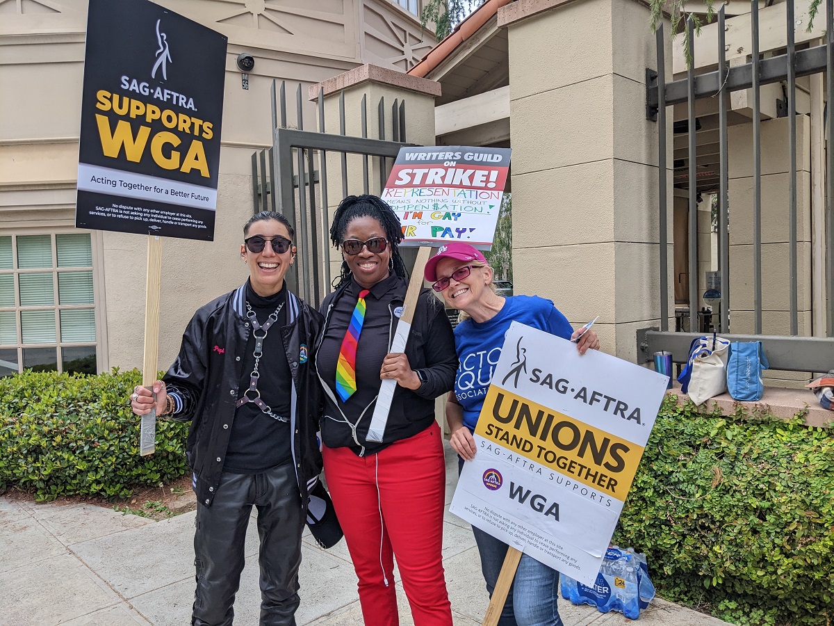 Image of Vico Ortiz and two women actors stand together smiling and posing with picket signs outside of Warner Bros. studios in Los Angeles during the WGA Strike. Vico is a non-binary Latine with short, dark, slicked-back hair wearing sunglasses, a black jacket over a black shirt and black pants, and a black leather harness around their chest. Their sign reads "SAG-AFTRA Supports WGA. Acting together for a better future. The woman in the center is Black and has long, black braids tied half-up, half-down. She's wearing a black hoodie over a black buttondown shirt and a rainbow tie with red pants. Her sign reads, "Writers Guild on Strike! Representation means nothing without Compen$ation! I'm Gay for Fair Pay!" The woman on the right is older and white. Her hair is in a ponytail and she's wearing a red LA Dodgers cap and a blue Actors Equity Association t-shirt. Her sign reads "SAG-AFTRA - Unions Stand Together! SAG-AFTRA supports WGA"