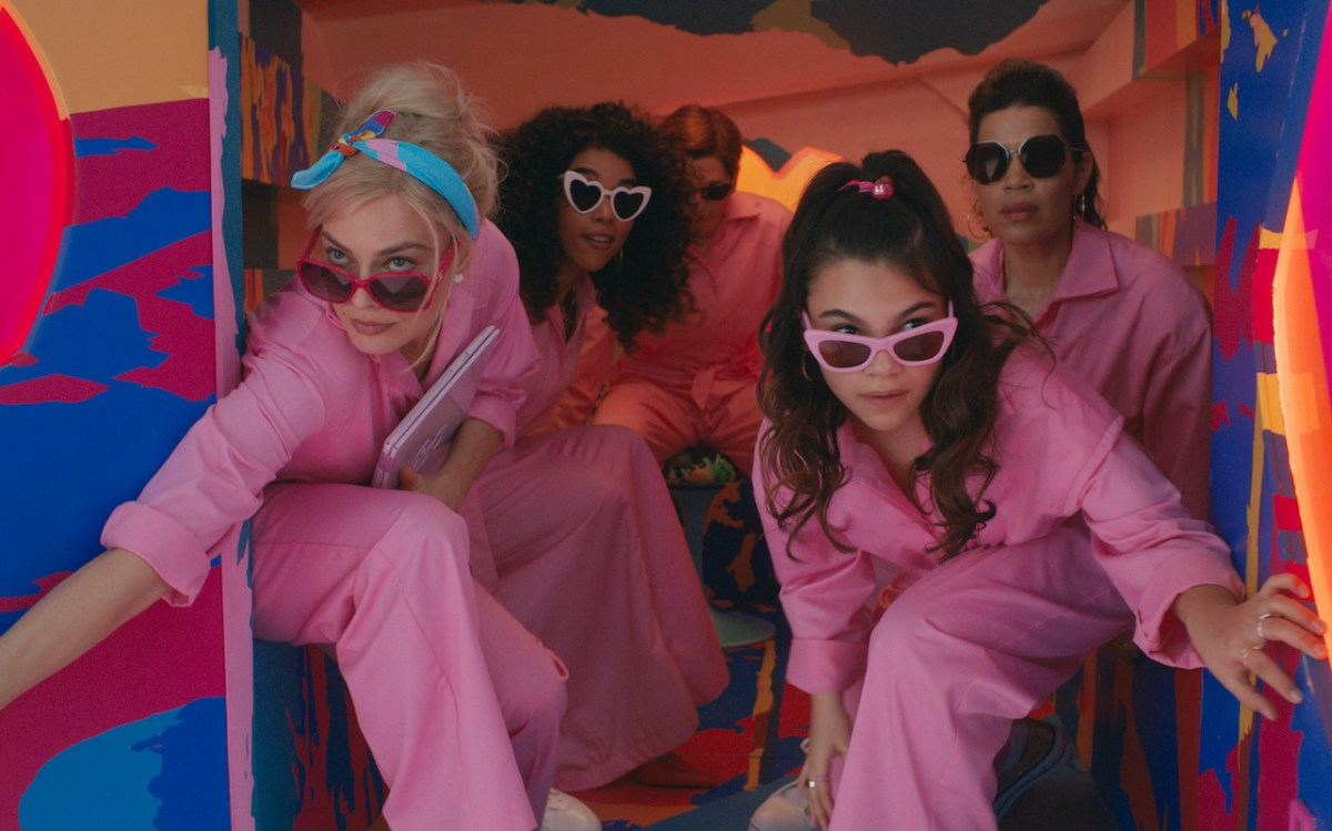Women dressed in pink jumpsuits and pink sunglasses lean out of the back of a van in a scene from Barbie.