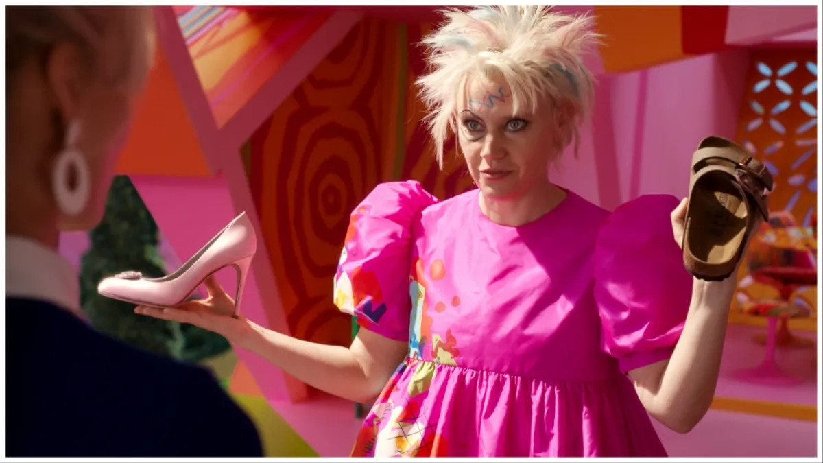 Kate McKinnon, in a pink dress with marker on her face and messily chopped-off hair, holds up a high heel and a birkenstock sandal in a still from 'Barbie'.