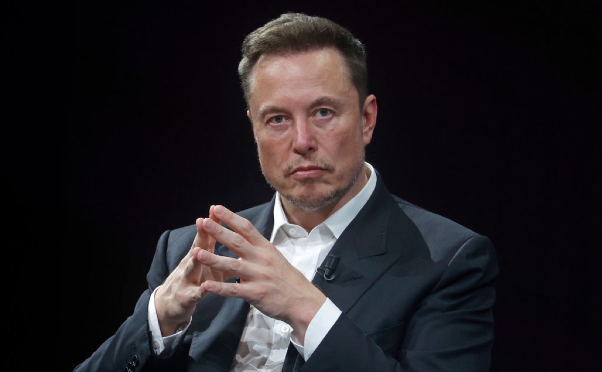 Elon Musk, alone against a black background, pressing his fingertips together and looking at the camera, displeased.