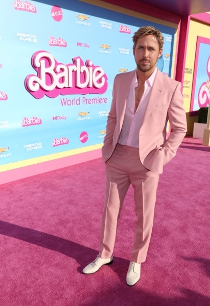 Ryan Gosling wears a light pink suit, white unbuttoned shirt, and white shoes on a pink carpet with the Barbie logo behind him.