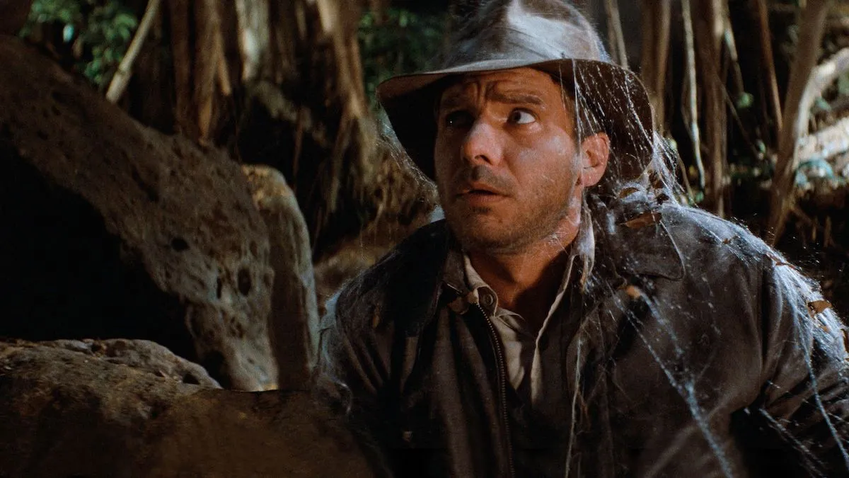 Rugged adventurer Indiana Jones looks perturbed while walking through cobwebs in a cave in "Raiders of the Lost Ark"