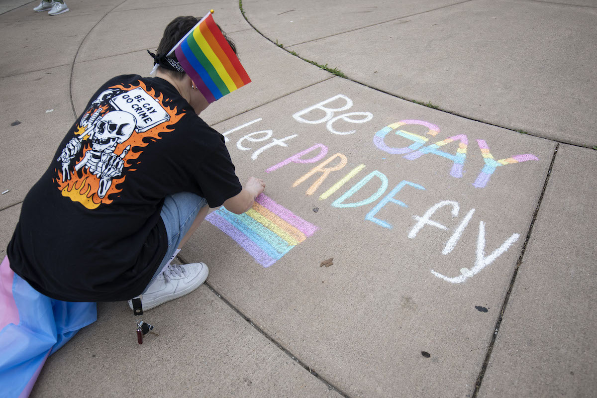A person crouches and draws a Pride message on the sidewalk in colorful chalk.