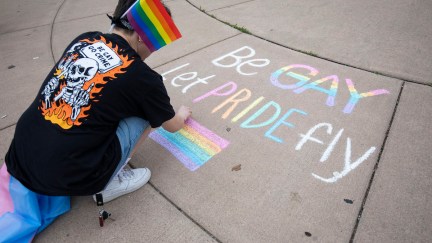 A person crouches and draws a Pride message on the sidewalk in colorful chalk.