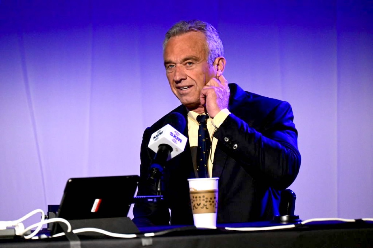 Robert F. Kennedy Jr. sitting at a microphone, grasping his earlobe with a look of uncertainty.