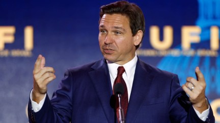 Ron DeSantis looks bewildered and points up with both hands while speaking into a microphone.