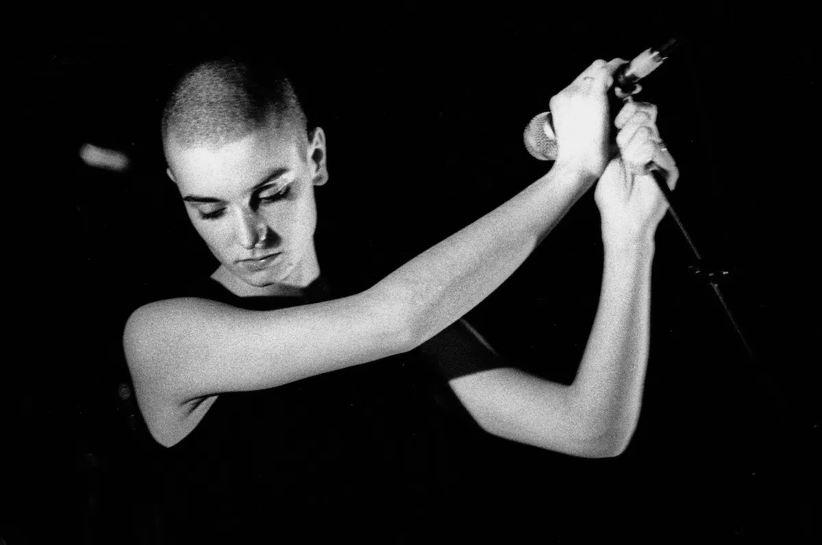 A black and white photo of a young Sinead O'Connor holding a microphone mournfully.