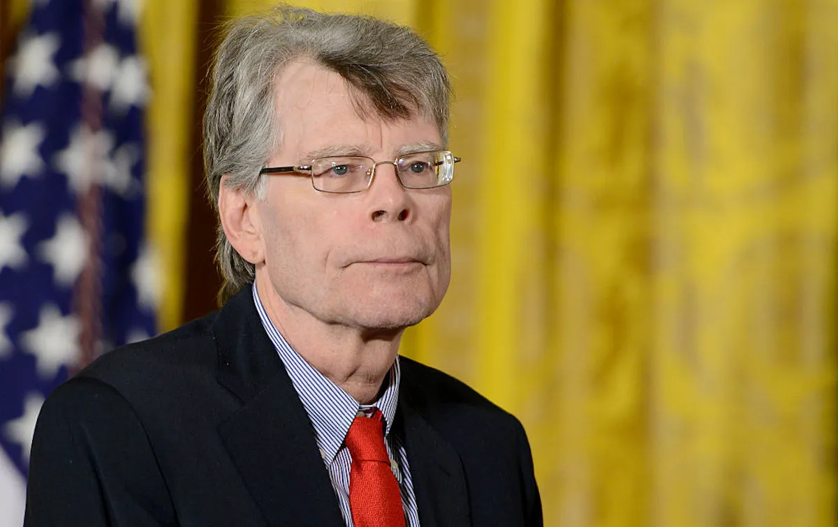 Stephen King looks off camera, wearing a red tie and standing in front of a yellow curtain and American flag.