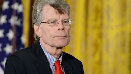 Stephen King looks off camera, wearing a red tie and standing in front of a yellow curtain and American flag.