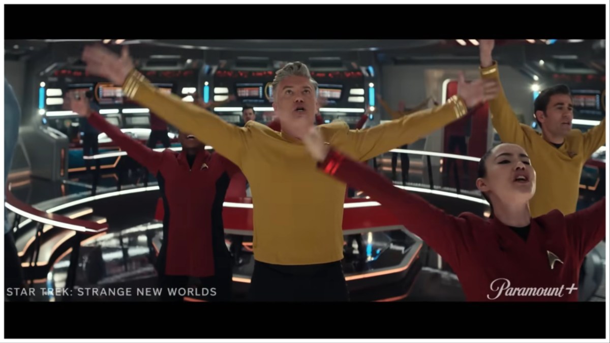Screengrab from the 'Strange New Worlds' musical episode, "Subspace Rhapsody." Captain Pike is in the center of several crew members on the bridge, all of whom have their arms in the air, mid-song.