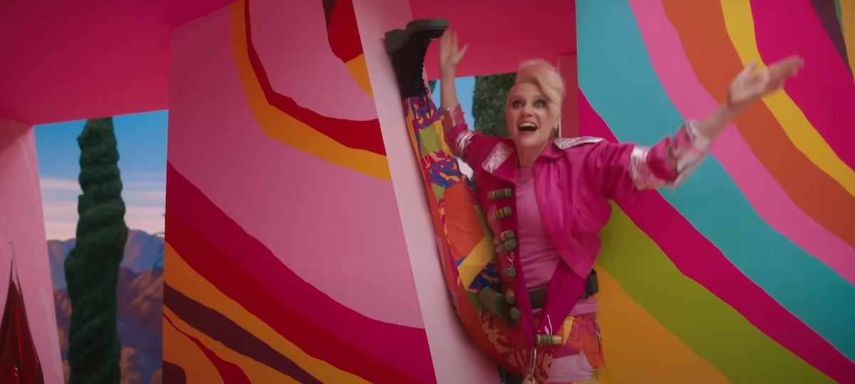 Kate McKinnon stands in a psychedelic-looking pink house, with short hair and weird makeup. One of her legs is up against the wall in a split.