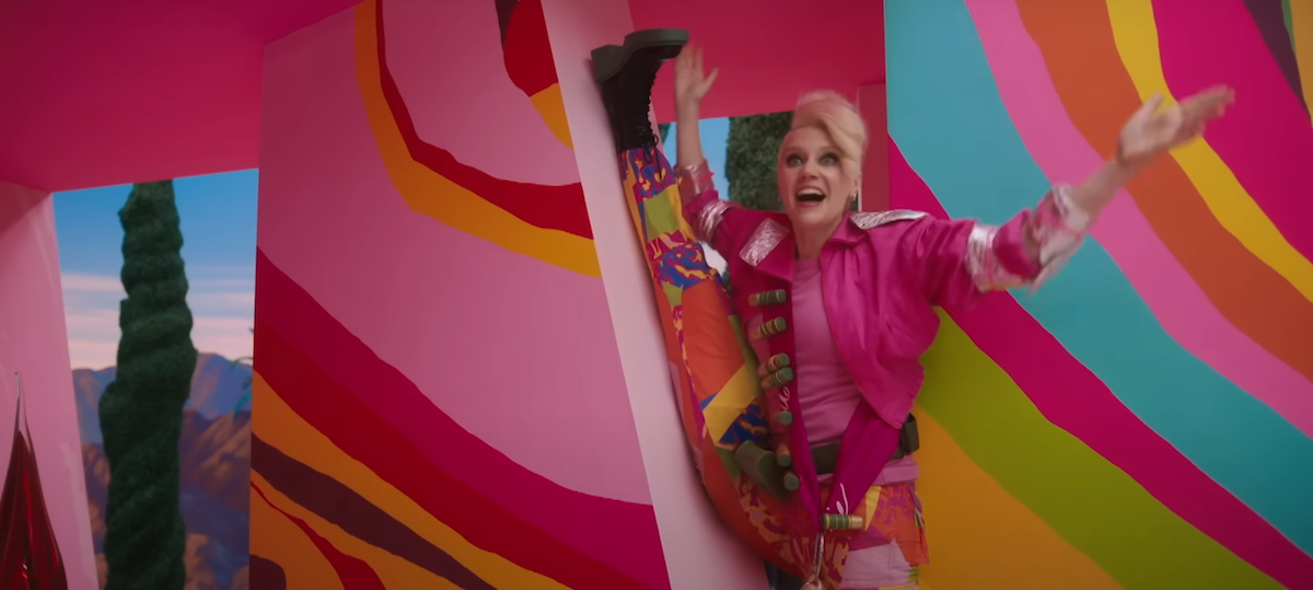 Kate McKinnon stands in a psychedelic-looking pink house, with short hair and weird makeup. One of her legs is up against the wall in a split.