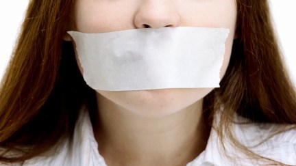 The bottom of a woman's face with a large piece of tape covering her mouth, silencing her.