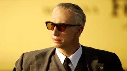 An older white man in a suit and large sunglasses stares into the distance in 