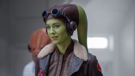 Mary Elizabeth Winstead as Hera Syndulla in a scene from Disney+'s 'Ahsoka.' Hera is a Twi'lek with green skin, bright blue eyes, and two long, tentacle-like appendages coming out of the back of her head and hanging behind her. She wears a brown leather aviator hat with goggles at her forehead and a brown leather bomber jacket with light brown fleece at the collar.