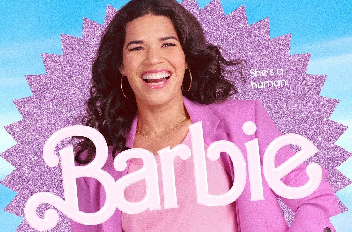 America Ferrera in her 'Barbie' movie poster. She is a Latina with long, wavy dark hair wearing a pink suit jacket over a lighter pink blouse and thin gold hoop earings. She's smiling a big smile as she stands in front of a pink glittery starburst against a blue backdrop. The text on the right reads "She's a human." and the title 'Barbie' in the Barbie font is across her chest.