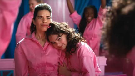 America Ferrera as Gloria and Ariana Greenblatt as Sasha in 'Barbie.' Both are brown Latinas. Gloria is a woman with long, straightened dark brown hair styled half up-half down. Sasha's hair is also long and dark brown, but much fuller with waves, and held half-up half-down with a hair tie that's pink little balls. Both are wearing long-sleeved, pink jumpsuits as Sasha leans her head on Gloria's shoulder.