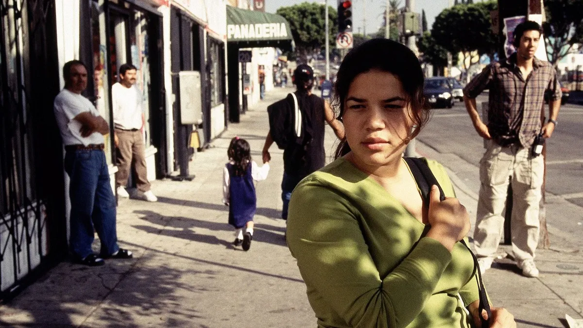 America Ferrera as Ana in 'Real Women Have Curves.' She is a Latina who is a high school senior walking down a Los Angeles street as she looks at something off in the distance. Her long, dark hair is in a ponytail with long tendrils hanging down one side of her face. She's wearing a green, v-necked long-sleeved shirt and holds one backpack strap on her shoulder with both hands.