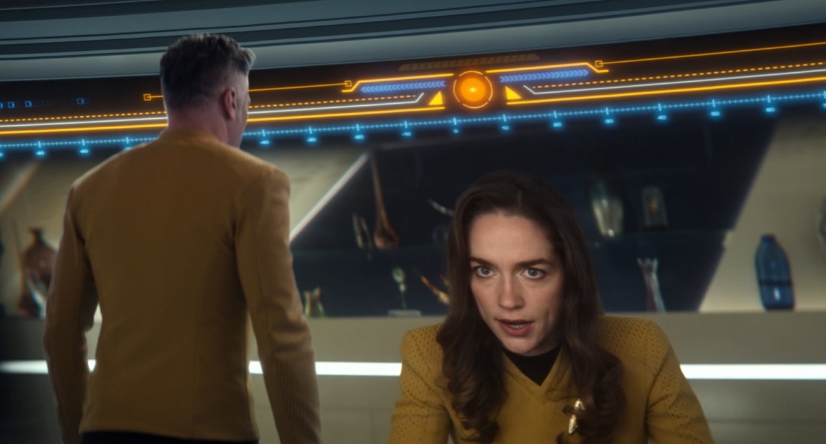 Image of Anson Mount as Captain Pike and Melanie Scrofano as Captain Batel in a scene from 'Star Trek: Strange New Worlds.' Pike is out of focus in the foreground with his back turned to the camera. We see Batel, a white woman with long, brown hair wearing a gold Starfleet uniform on the large bridge viewscreen. She looks upset.