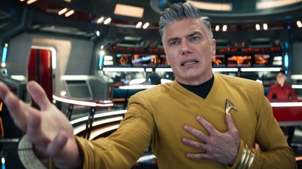 Image of Anson Mount as Captain Christopher Pike in a scene from 'Star Trek: Strange New Worlds.' He is a white man with perfectly coiffed salt-and-pepper hair wearing a gold Starfleet uniform. He's on the bridge of the Enterprise singing on his knees with one hand on his chest and the other outstretched.